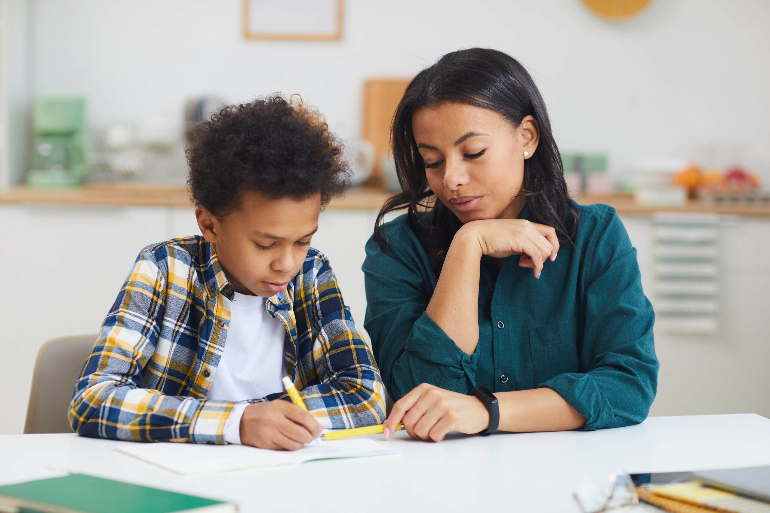 Portrait of young African-American boy studying with female teacher watching him in class,